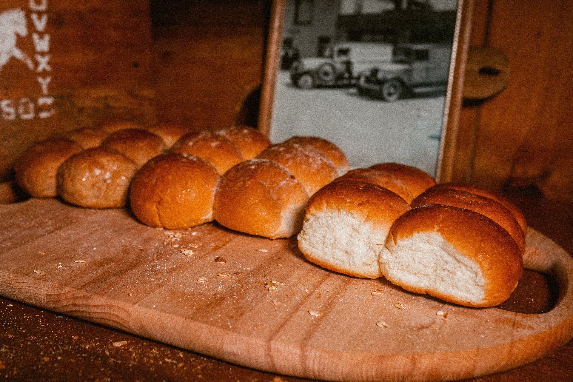 Golden-brown dinner rolls arranged on a wooden cutting board, with a framed vintage photograph of cars in the background.