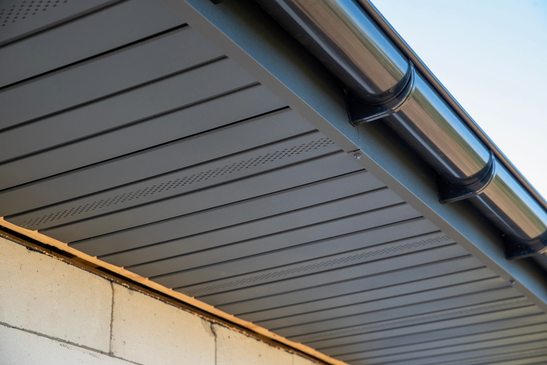 Dark gray vented soffit panels installed under a roof eave, adjacent to a light-colored cinder block wall and gutter. Dark gray vented soffit panels installed under a roof eave, adjacent to a light-colored cinder block wall and gutter.