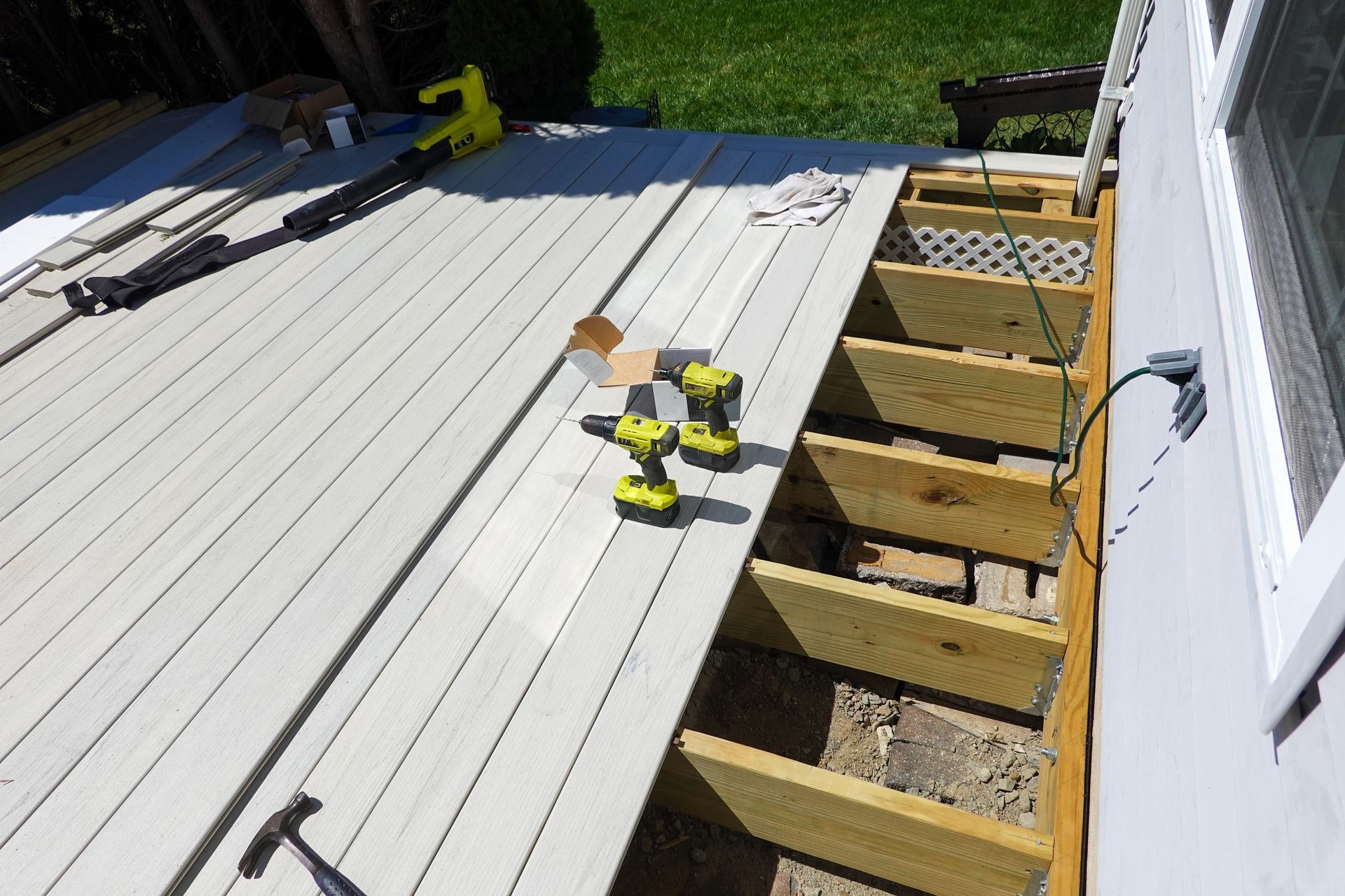 A construction scene of a partially laid light-colored deck against a building, featuring tools and wooden joists. A construction scene of a partially laid light-colored deck against a building, featuring tools and wooden joists.