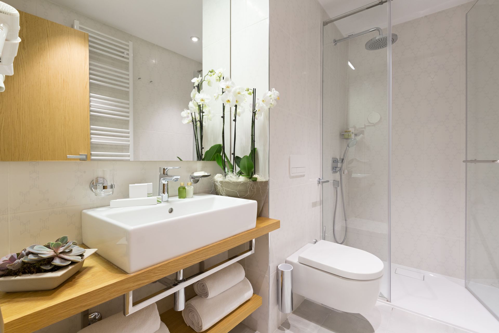 A bright, modern bathroom featuring a white vessel sink on a wooden vanity, a walk-in glass shower, and a white toilet. A bright, modern bathroom featuring a white vessel sink on a wooden vanity, a walk-in glass shower, and a white toilet.