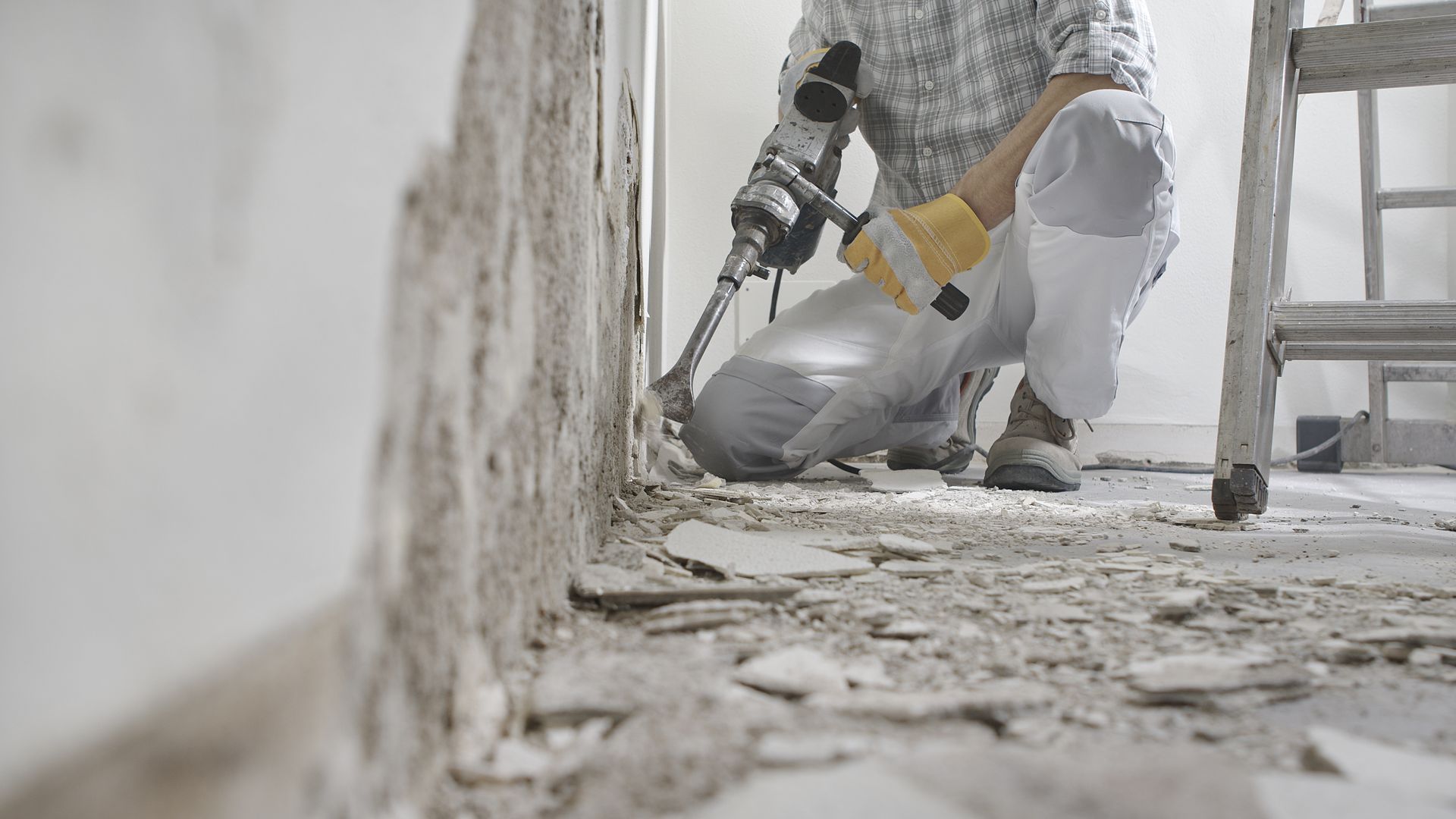 A person in work clothes uses a power tool to remove plaster from a wall in a room under construction. A person in work clothes uses a power tool to remove plaster from a wall in a room under construction.