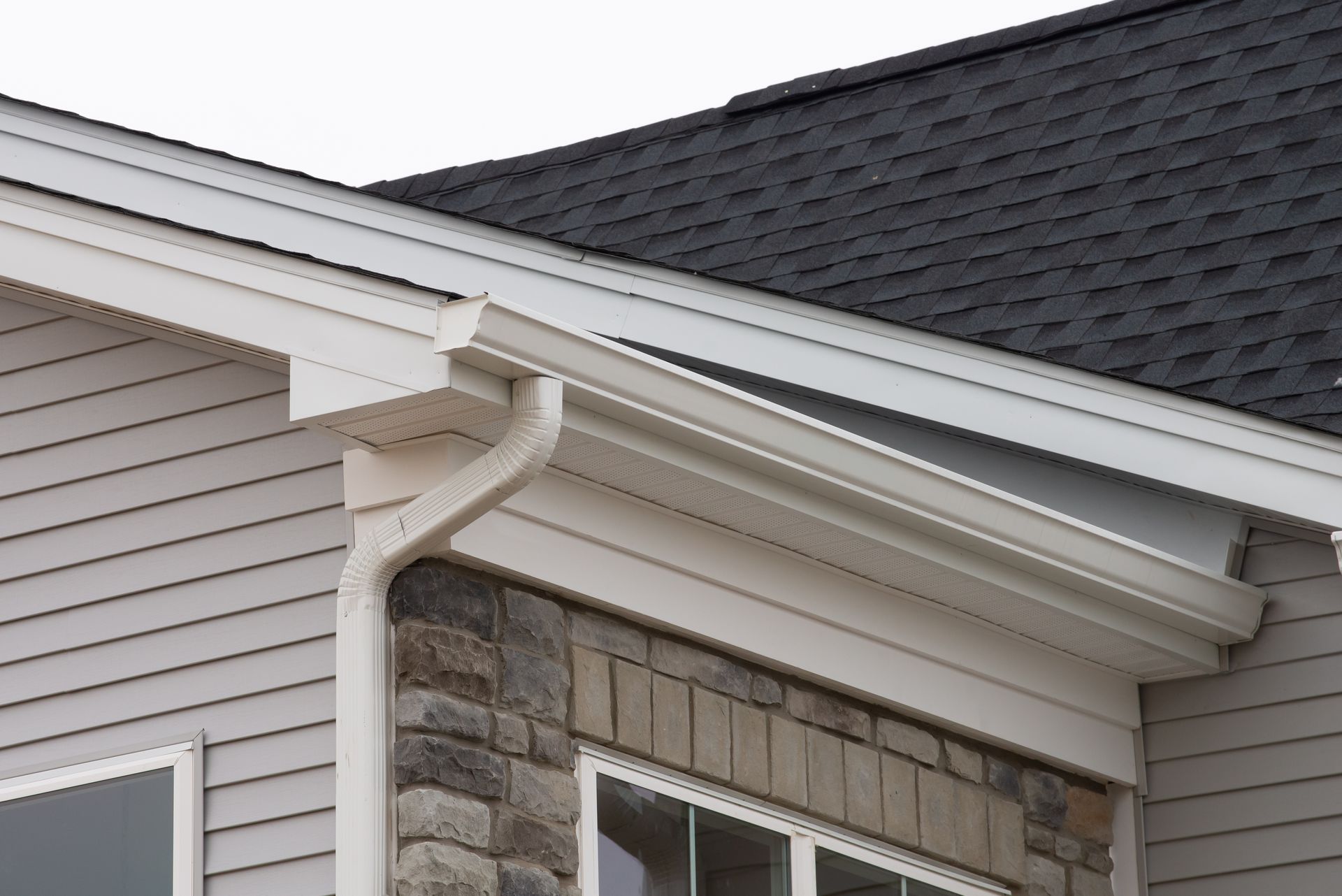 White rain gutters and a downspout attached to the corner of a house with stone accents and grey vinyl siding. White rain gutters and a downspout attached to the corner of a house with stone accents and grey vinyl siding.