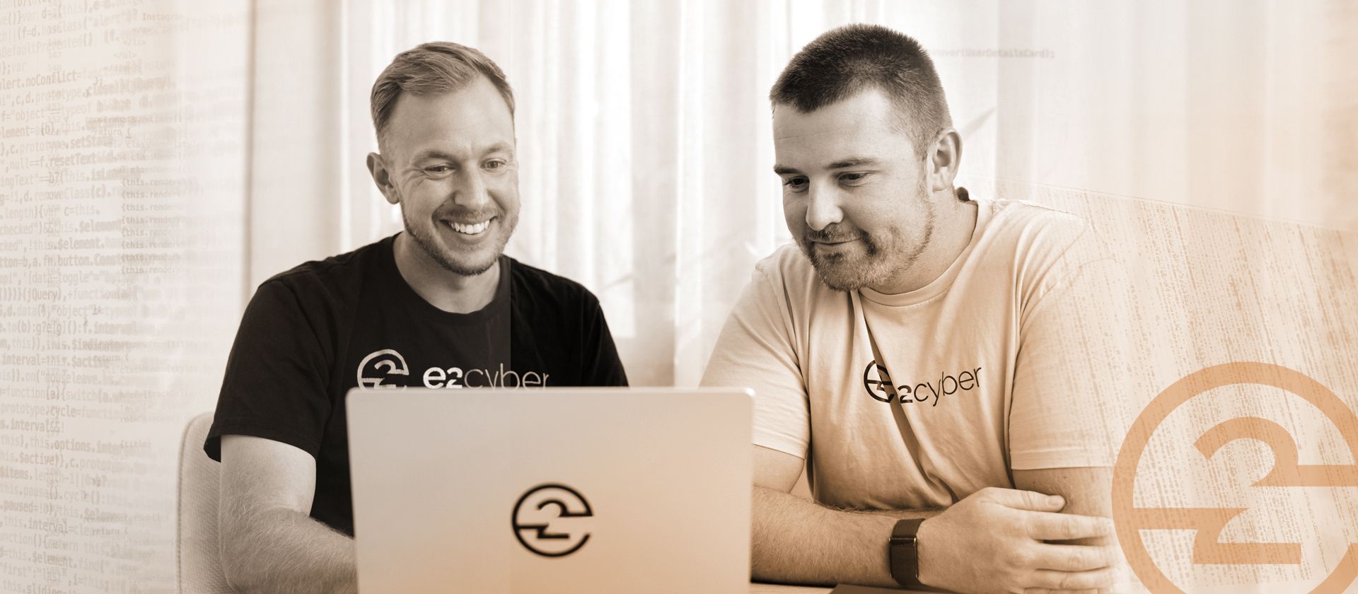 Two colleagues in branded shirts look at a laptop screen together in a softly lit, professional office environment.