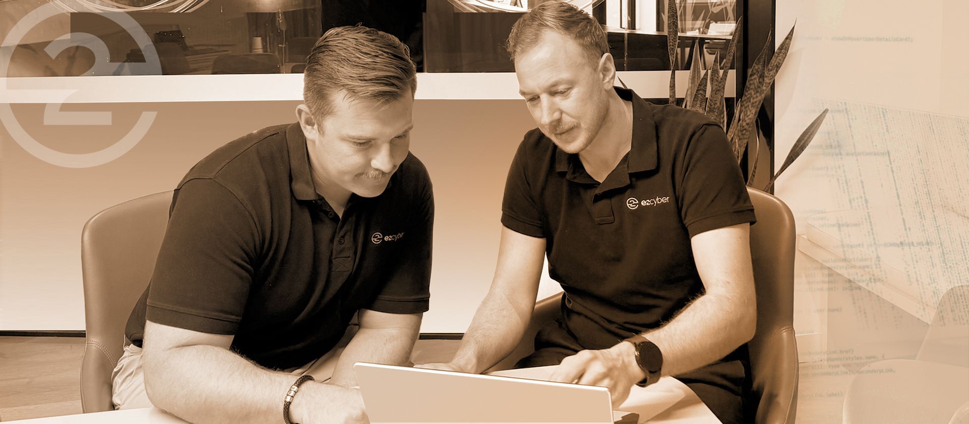Two men in black polo shirts review plans together at a table in an office-like setting.