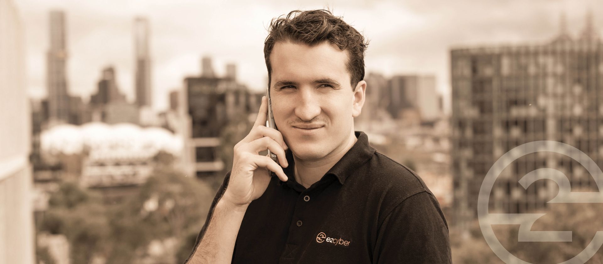 Man on a phone in a cityscape, wearing a black polo shirt and smiling at the camera.