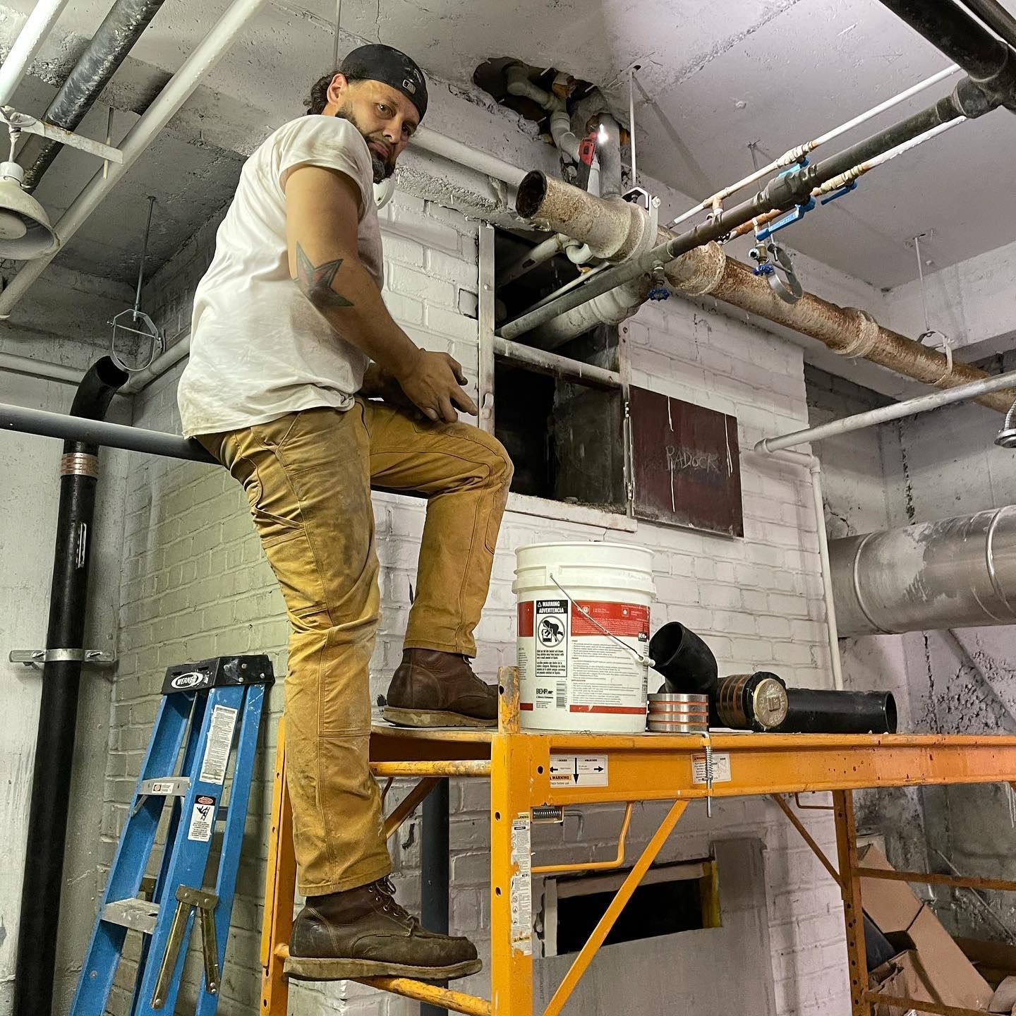 A man is sitting on a ladder in a room with pipes.