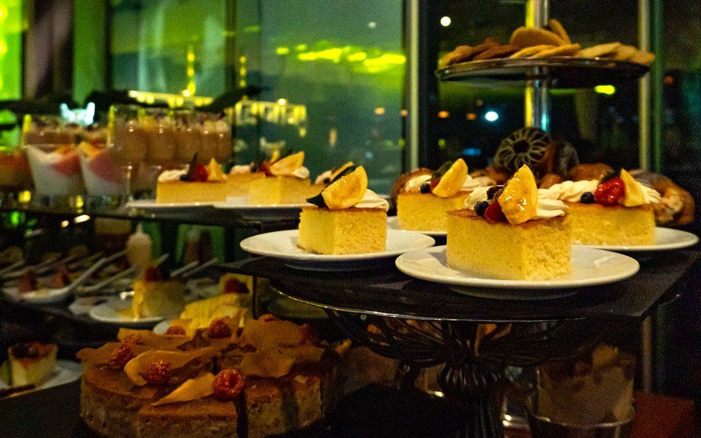 Bufé de postres con tartas en capas, galletas y otros dulces en bandejas y soportes escalonados, con poca iluminación.