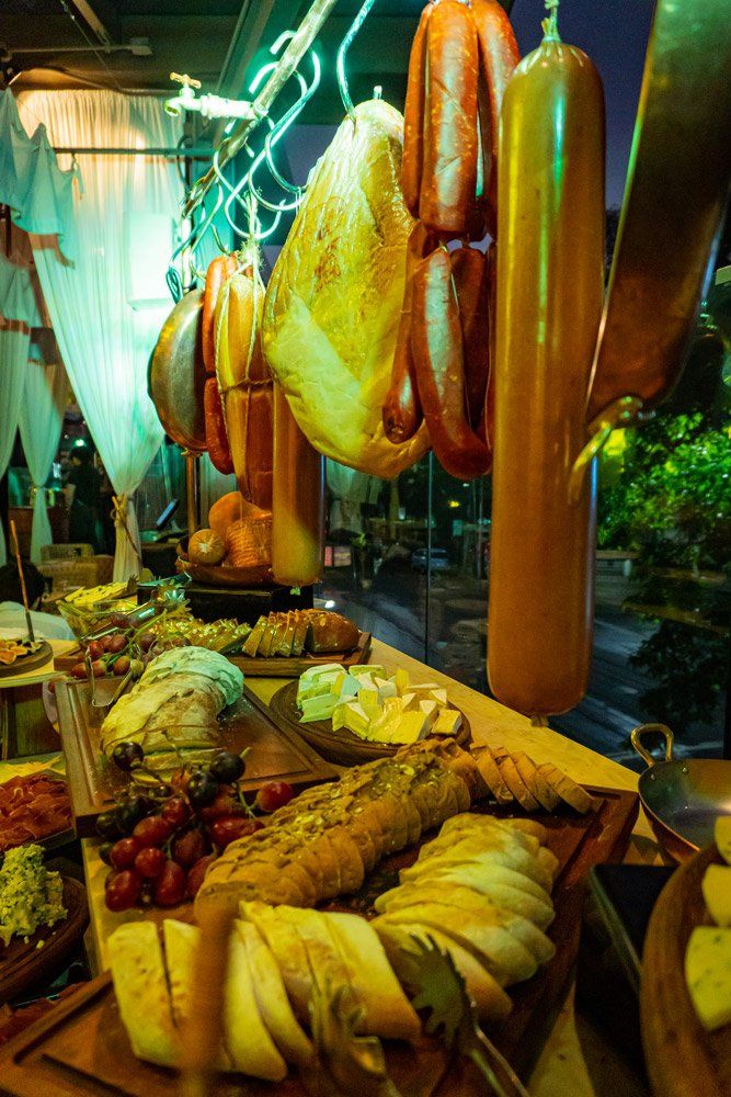 Exhibición de comida: pan de molde, queso, embutidos colgados de una barandilla, mesa buffet con platos variados.