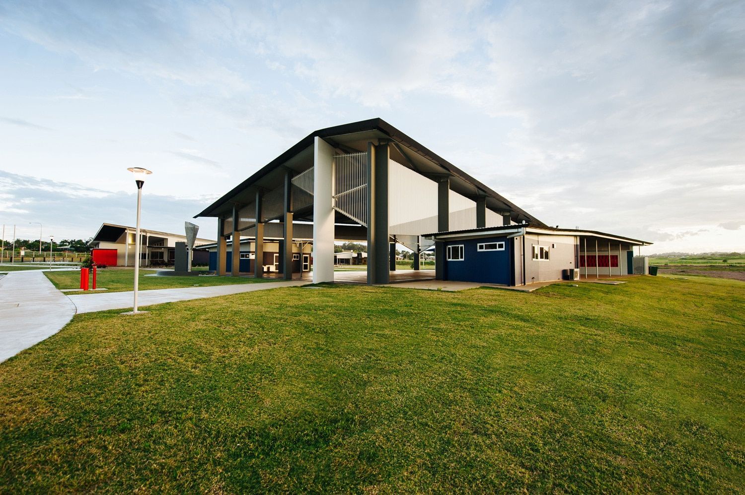 A Large, Modern Building With a Sloping Roof and Open Sides on a Grassy Hill Under a Cloudy Sky — BOLD Architecture + Interior Design In Mackay, QLD