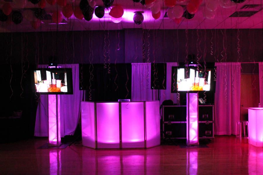 DJ setup with pink lighting, monitors, and draped backdrop, balloons overhead.