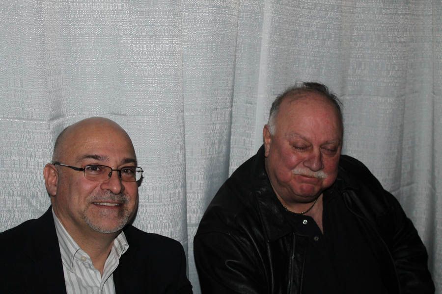 Two men seated next to each other in front of a light-colored backdrop. One man wears glasses, the other a black jacket.