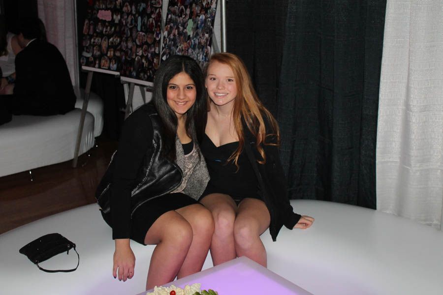 Two smiling women in black dresses sit together on a white platform.