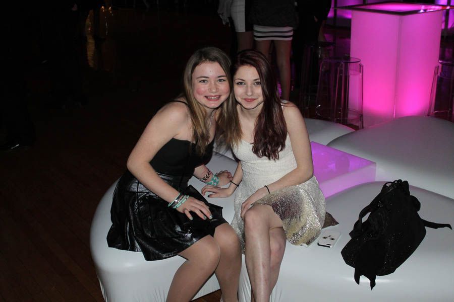Two young women smiling, sitting on a white curved sofa in a dimly lit space with a pink glowing bar.