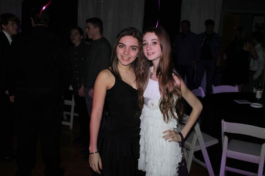 Two young women posing at a party. One in a black dress, the other in white fringe. Dark background.