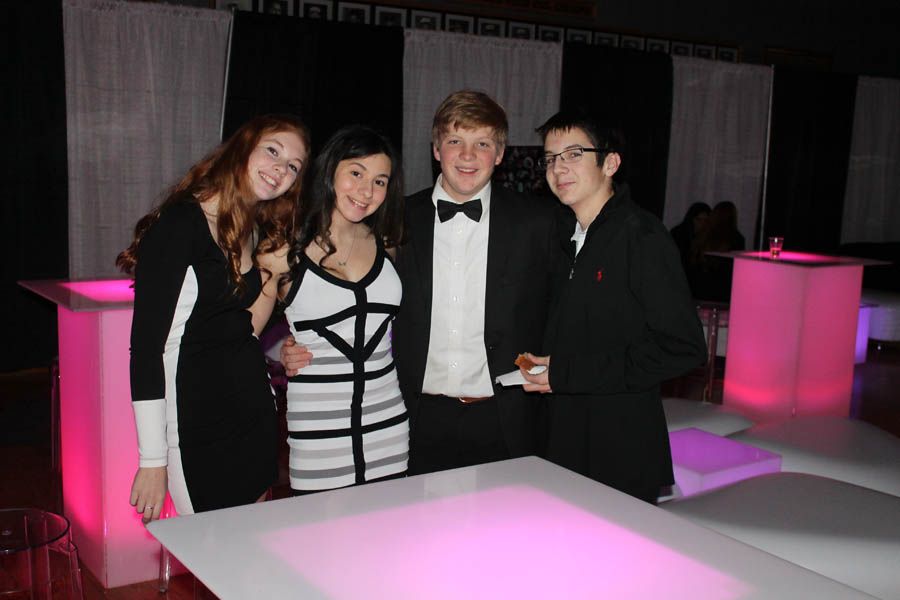 Four young people at a formal event, posing by a glowing table. One woman wears a black and white dress, others in tuxedos.