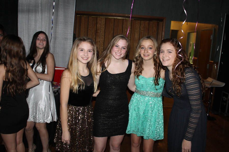 Four smiling girls in party dresses, arms linked, posing in a room.