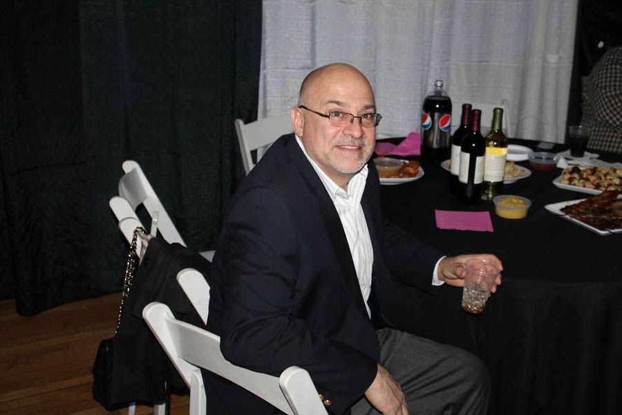 Man sitting at a table with drinks and food, wearing a blazer, smiling indoors.