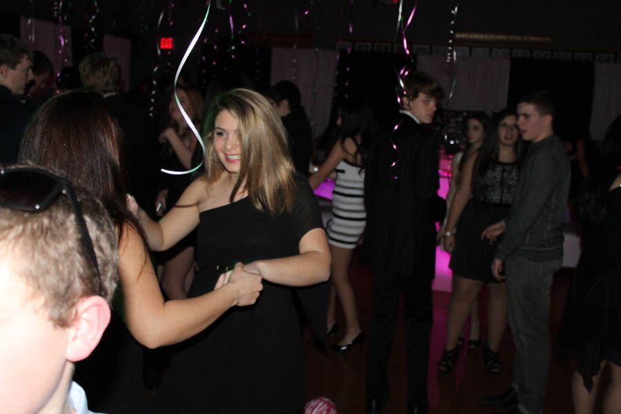 People dancing at a party with black dresses and suits. Pink and white lights illuminate the dance floor.