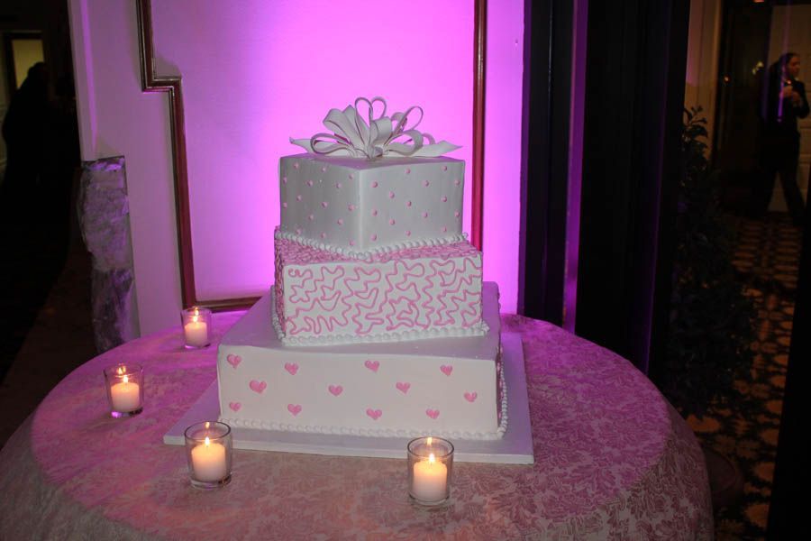 Three-tiered cake decorated with pink hearts, bows, and patterns, lit by pink uplighting, on a round table.