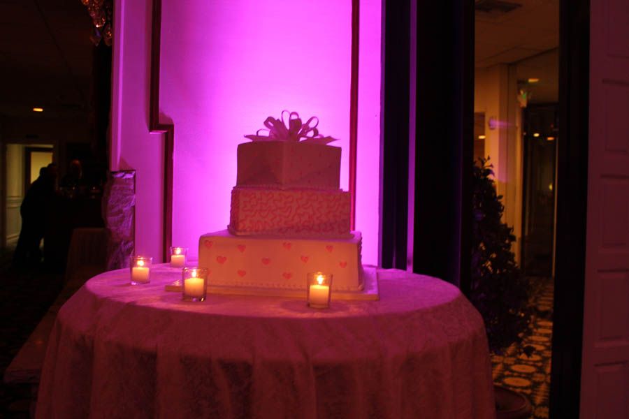 Three-tiered cake with pink hearts and a bow on a round table, lit by candles and pink light.