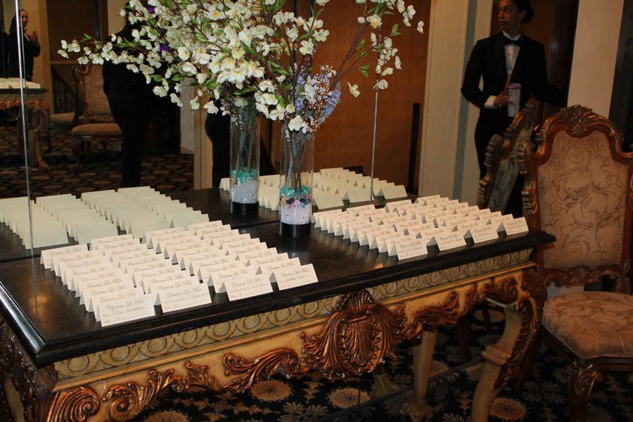 Wedding place cards on ornate table with flower arrangements and a server.