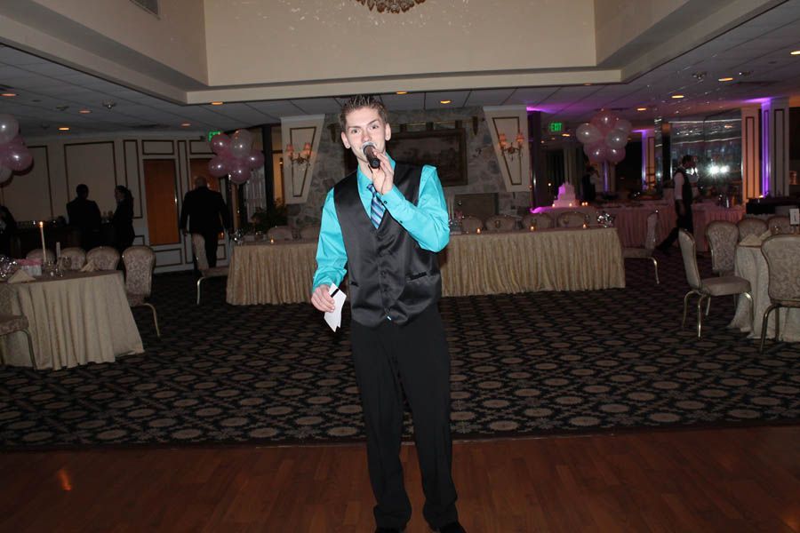 Man in teal shirt and vest speaks into a microphone at a formal event.