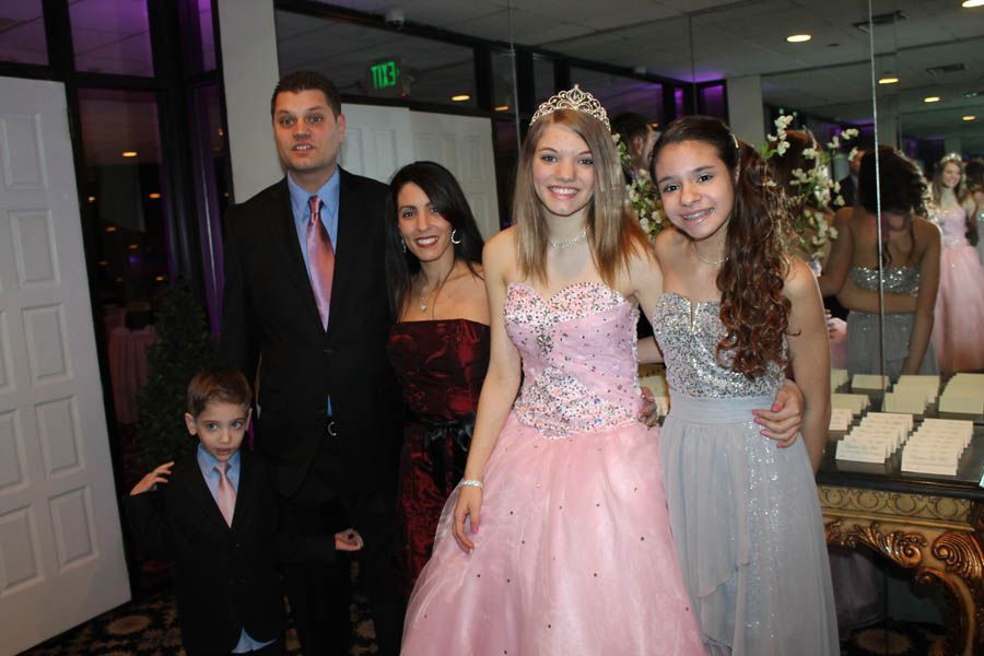 Family at an event: man, woman, two girls in dresses, and boy in a suit pose near a table and a mirror.