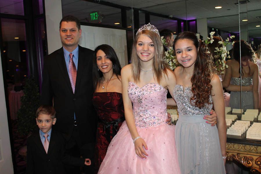 Family poses at an event; a girl in a pink gown with a tiara is flanked by another girl in silver, parents, and a young boy.