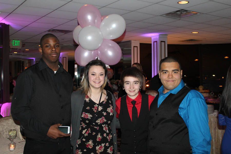 Four people pose for a photo indoors, near pink and white balloons. One wears a black suit, and another a floral dress.