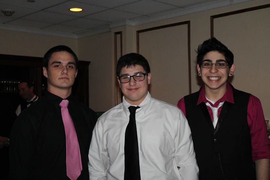 Three young men in formal attire, smiling, indoors. One in pink tie, one in black tie, one in a vest and red shirt.