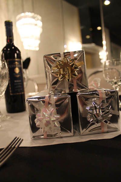 Three wrapped gifts stacked on a table with a wine bottle, bow details in gold, silver, and pink.