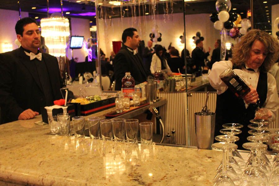 Bartenders preparing drinks at a formal event; champagne flutes stacked; people in the background.