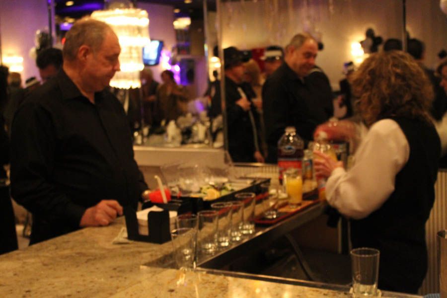 Man at bar watches bartender prepare drink at an event; other people in the background.