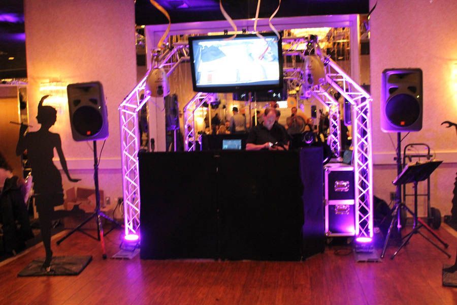 DJ setup with purple lighting, speakers, and a screen in front of a bar.