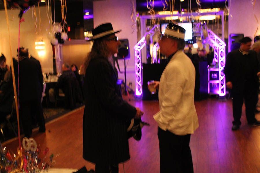 Two men talking at a party. One in black hat and coat, the other in white jacket, near a lighted structure.