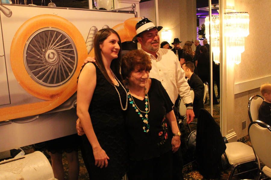 Three people pose for a photo in front of a car backdrop; woman on left, older couple in center.