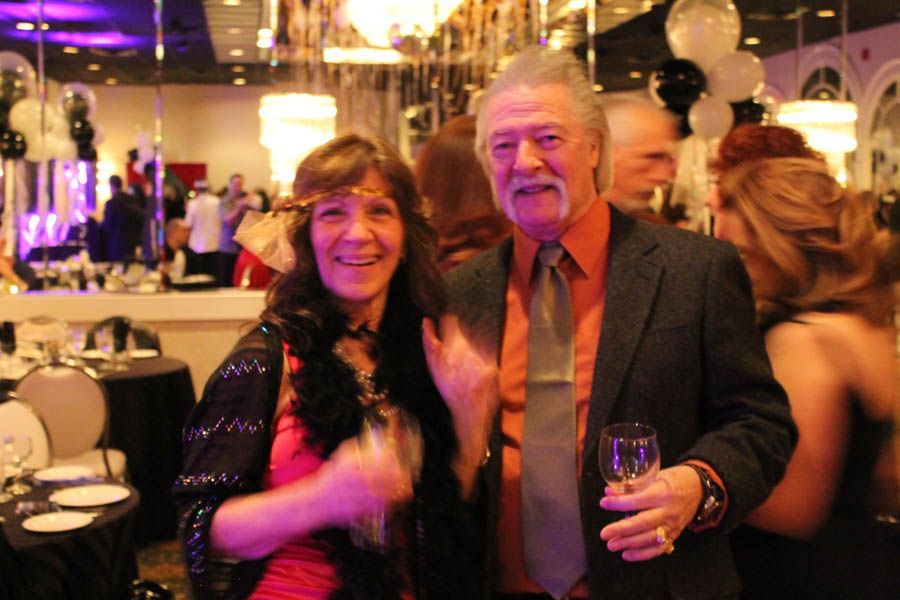 Couple at a party, woman with headband, man in suit holding a glass. Mirrors and decorations in the background.