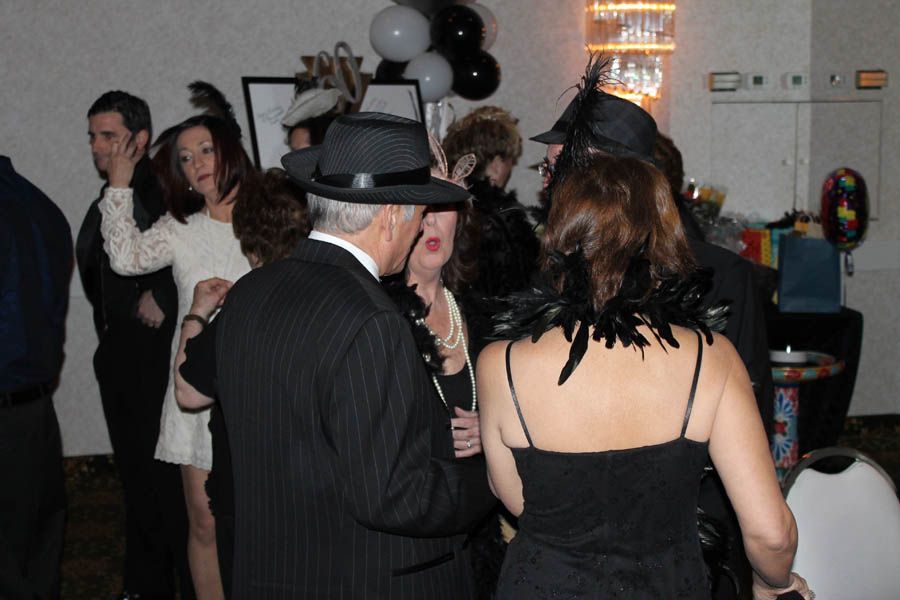 People in formal attire at a party, including a man in a pinstripe suit and fedora. Black and white balloons.