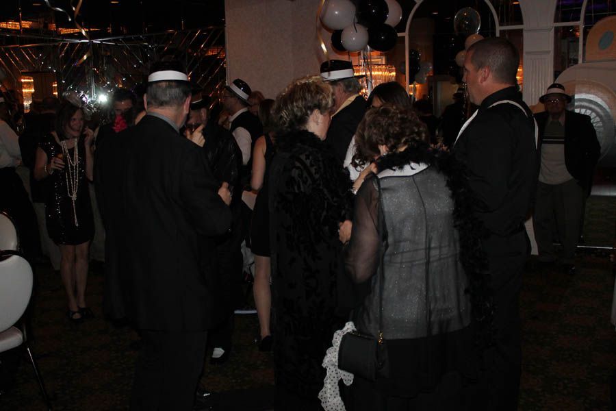 People in formal black attire at a gathering, some wearing hats and feather boas, with black and white balloons.
