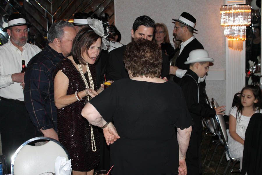 People at a party, some wearing hats and long necklaces. Inside, bright lighting, round tables.
