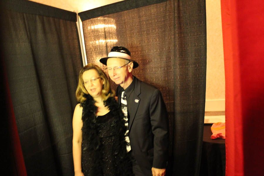 Couple posing in a photo booth. Woman in black dress and boa, man in suit and hat. Black curtain backdrop.