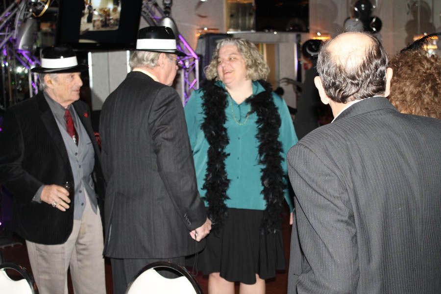 People at a party, some wearing hats and a woman with a boa. Inside, potentially a ballroom setting.
