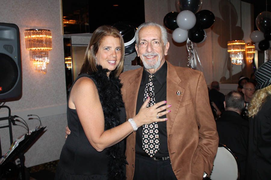 Woman with black boa and older man in suit pose at event with black and white balloons.