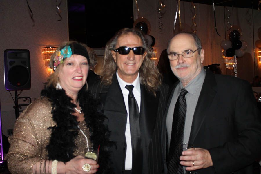 Three people at a party; woman in gold and black boa, man in sunglasses, man in a suit, smiles, indoors.