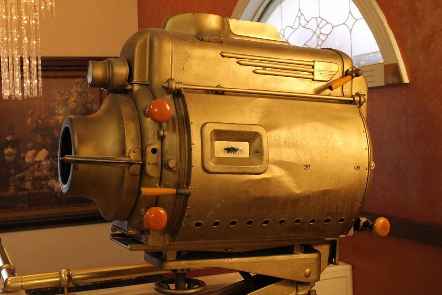 Old, gold-colored movie projector on a stand. Orange knobs and small window on front, near a window and wall.