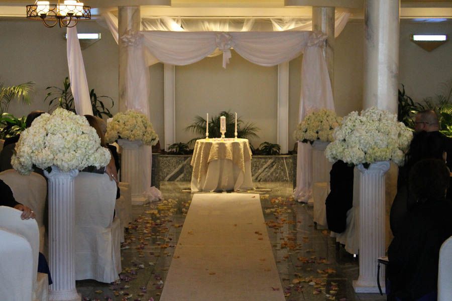 Wedding ceremony setup: white flowers atop pillars line a tan aisle, leading to draped altar and candlelit table.