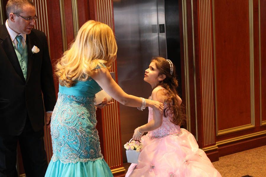 Woman in blue dress comforts girl in pink dress near elevator; man in suit watches.