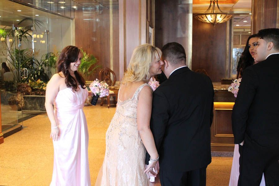 Wedding party indoors: Bride and groom embrace, others watch. Pale pink and gold tones.