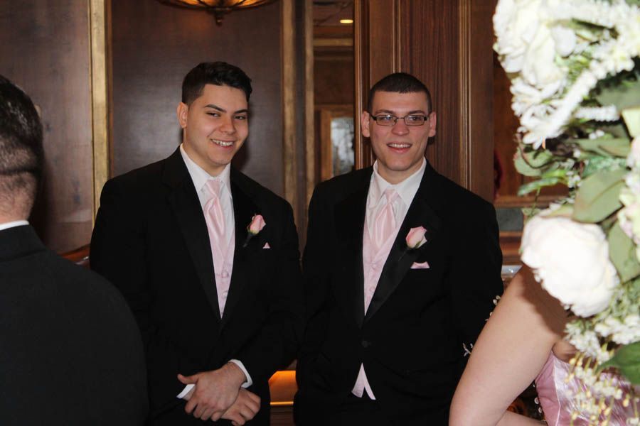 Two men in black suits and pink ties smiling at a wedding, near floral decorations.