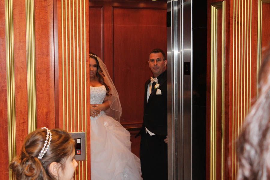 Bride and groom in wedding attire inside an elevator. The groom looks on, while the bride stands with her veil.